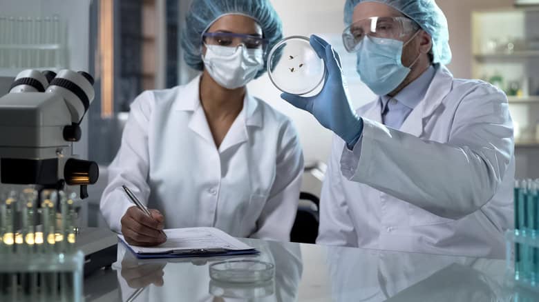 Two scientists in lab coats, masks, and gloves examine a petri dish. One holds the dish while the other takes notes on a clipboard. Laboratory equipment is visible in the background.