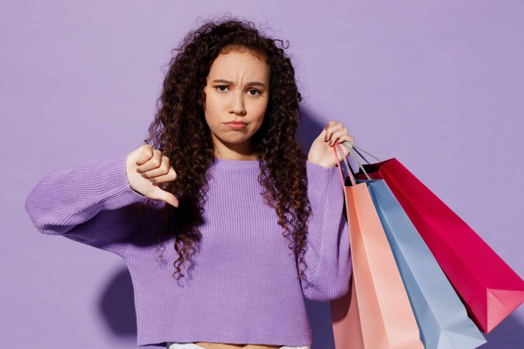A woman in a purple sweater shows a thumbs-down gesture while holding multiple shopping bags against a light purple background.