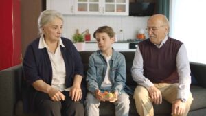Two elderly individuals and a young boy sit on a sofa. The elderly woman is speaking, the boy looks thoughtful while holding a bowl, and the elderly man is looking at the boy. A kitchen is in the background.