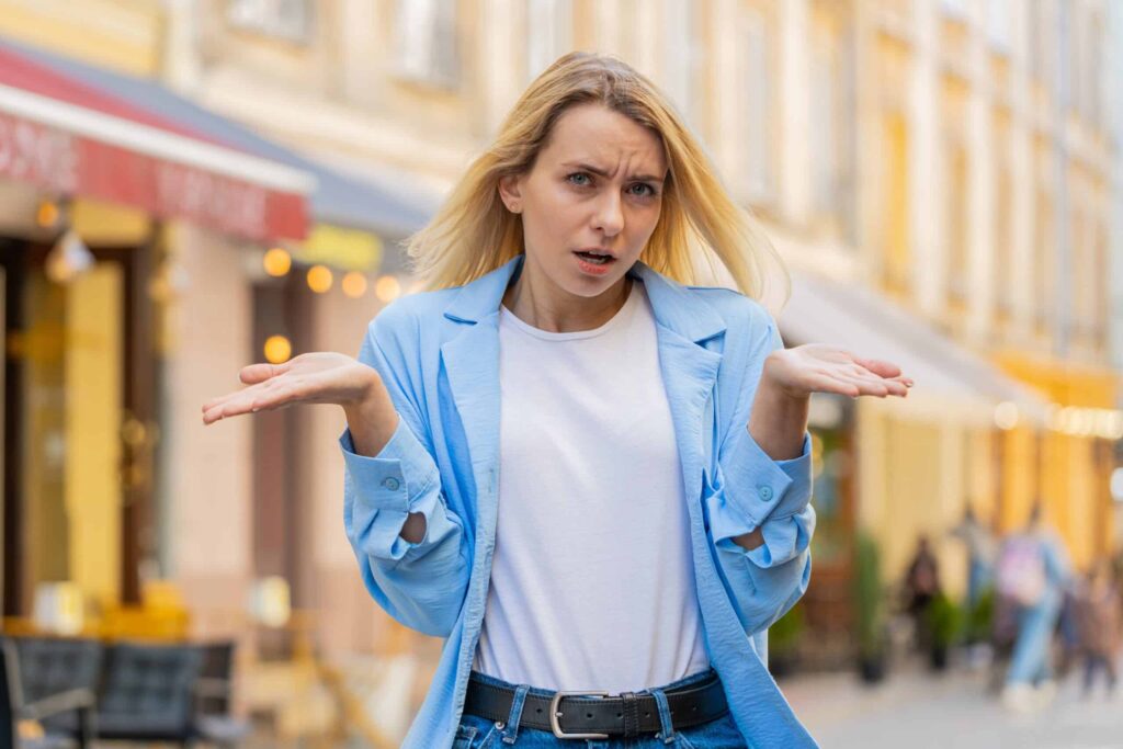 A woman with long blonde hair and a blue jacket is outdoors, standing with her arms outstretched and palms facing up, looking confused or questioning. There are shops and people in the background.