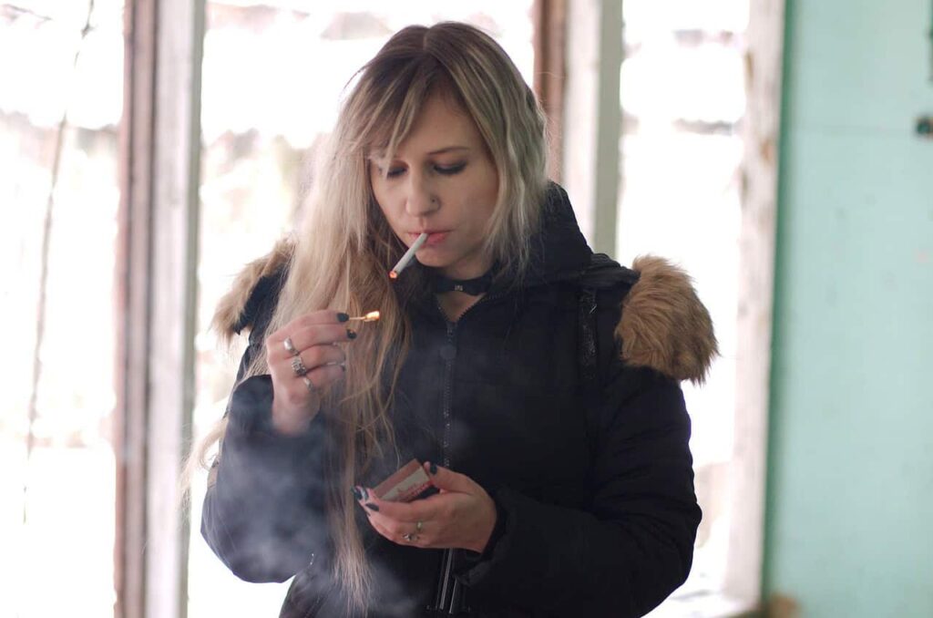 A woman with long hair standing indoors lights a cigarette using a match, holding a matchbox in her other hand. She is wearing a black jacket with a fur-trimmed hood.