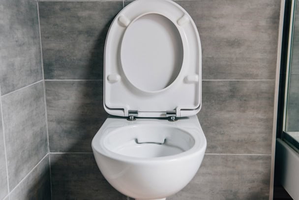 A white toilet with the lid up is mounted on a gray-tiled wall in a bathroom.