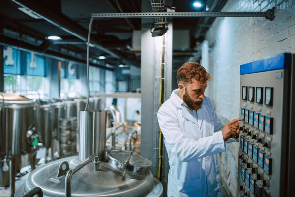 A man in a lab coat operates a control panel in a modern industrial facility with large stainless steel tanks.