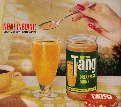 A hand holding a spoon with powder above a glass of orange drink next to an open jar of Tang Breakfast Drink and a lid on a table. A cup of coffee is in the background.