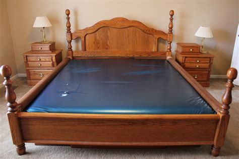 A wooden bed with a blue water mattress, flanked by two wooden nightstands each with a lamp, in a beige-colored room.