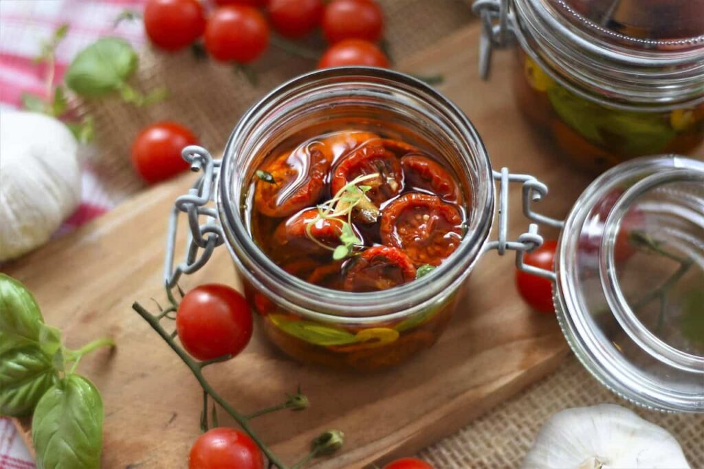 A glass jar filled with preserved sun-dried tomatoes in oil, topped with a sprig of herbs. Around the jar are fresh tomatoes, garlic, basil, and another open jar.