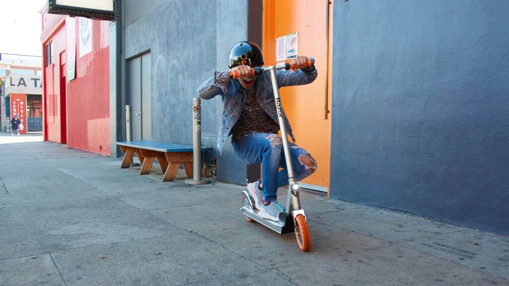 A person wearing a helmet rides an electric scooter near a colorful building while smiling and gripping the handlebars tightly.