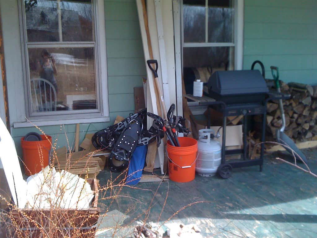 A porch with various items including a stack of wood, a grill, two buckets, a propane tank, some boards, a shovel, and a set of snowshoes.