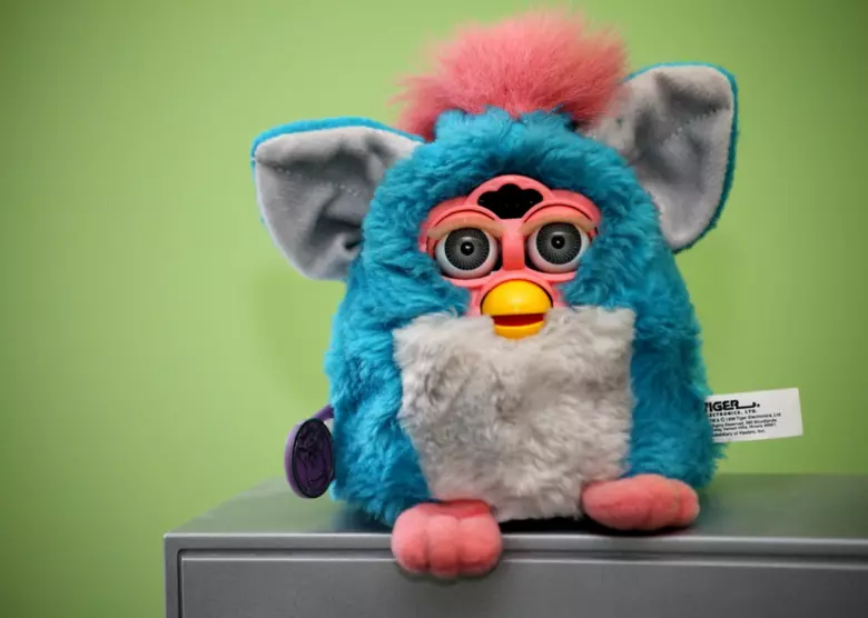 A blue and pink Furby toy with large ears and wide eyes sits on top of a gray surface in front of a green background.