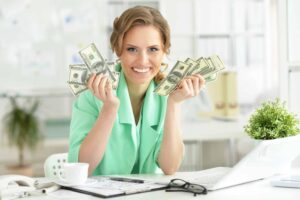 A woman in a green blazer smiles while holding stacks of US dollar bills in both hands in an office setting.
