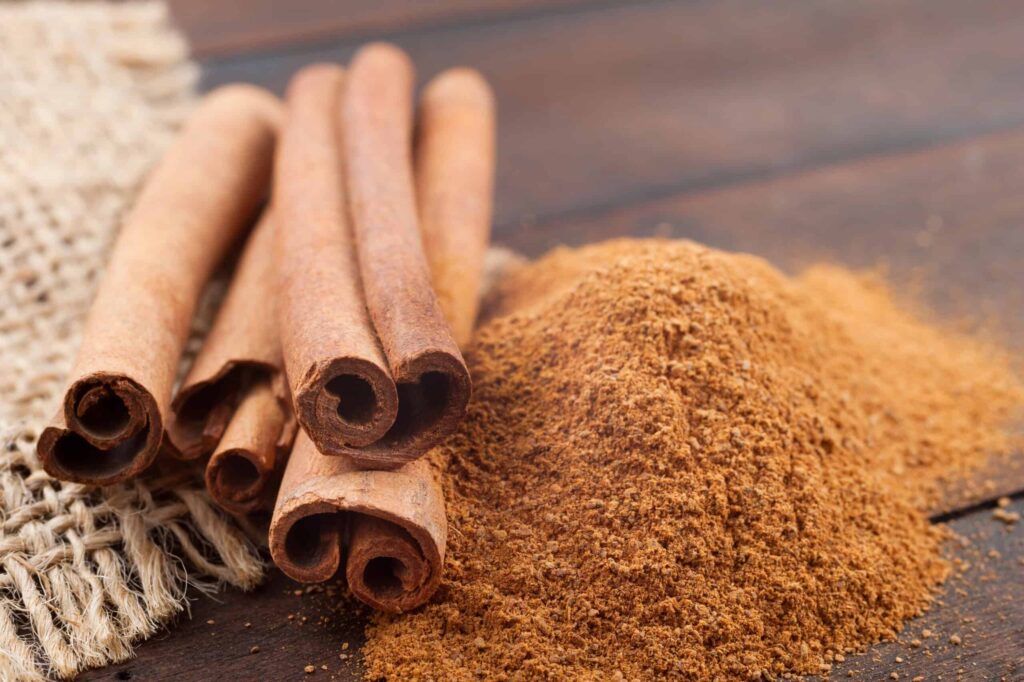 Cinnamon sticks lie next to a heap of ground cinnamon on a wooden surface with a piece of burlap fabric on the side.
