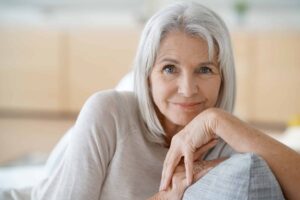 An elderly woman with white hair and a light-colored top sits and smiles while resting her hands on her chin.