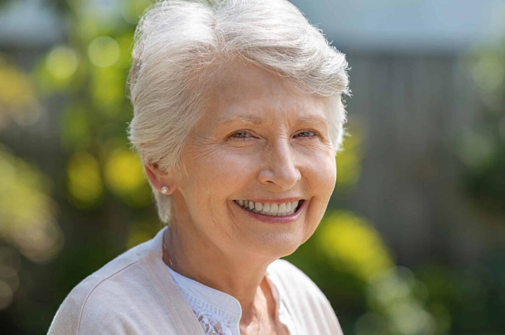 An elderly woman with short white hair smiles at the camera while standing outdoors.