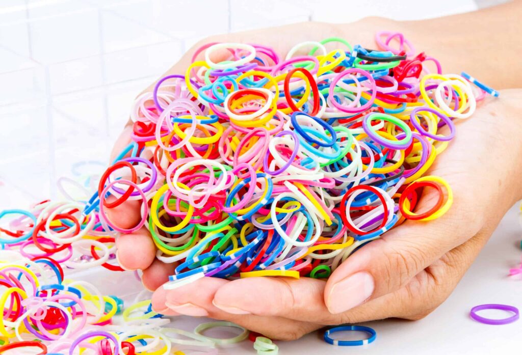 Two hands holding a large pile of multicolored rubber bands with more rubber bands scattered below.