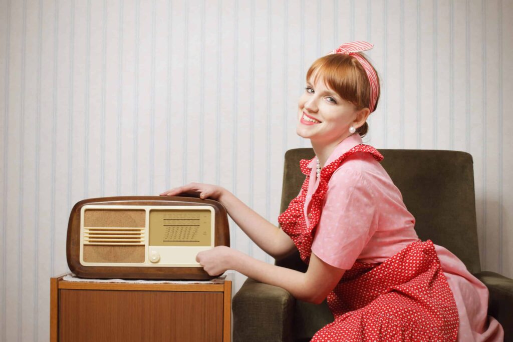 A woman in a pink and red polka dot outfit smiles while sitting next to and touching an old-fashioned radio on a small wooden table.