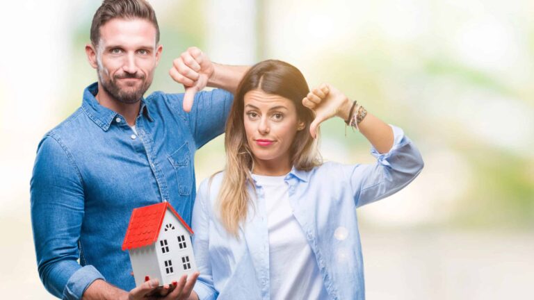 A man and a woman, both in denim shirts, hold a small house model. Both are giving thumbs-down gestures, suggesting dissatisfaction. The background is bright and blurred.