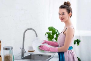 A woman wearing pink gloves and an apron is washing a plate at a kitchen sink. Plants and kitchen items are visible in the background.