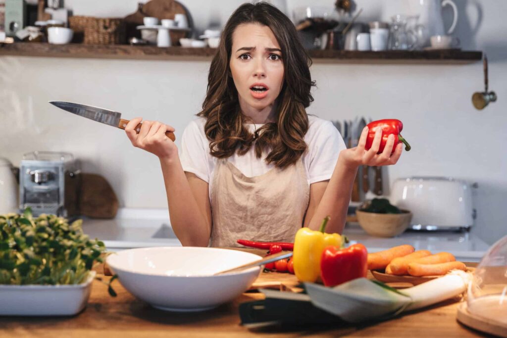 A woman in an apron stands in a kitchen holding a knife and a red bell pepper, looking confused. Various vegetables and kitchenware are spread out on the counter in front of her.
