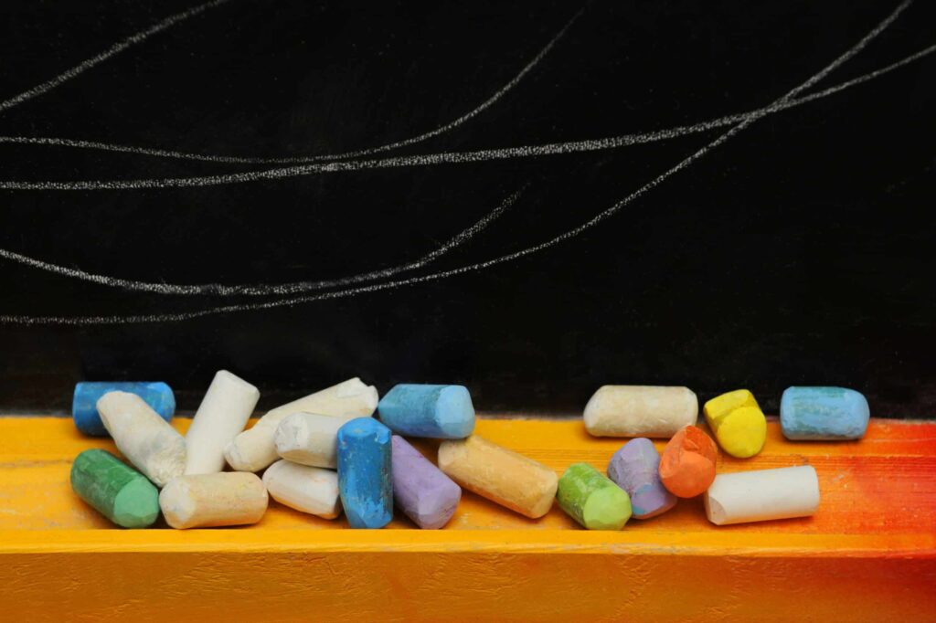 Pieces of multicolored chalk are arranged on a yellow chalk tray below a blackboard with white chalk markings.