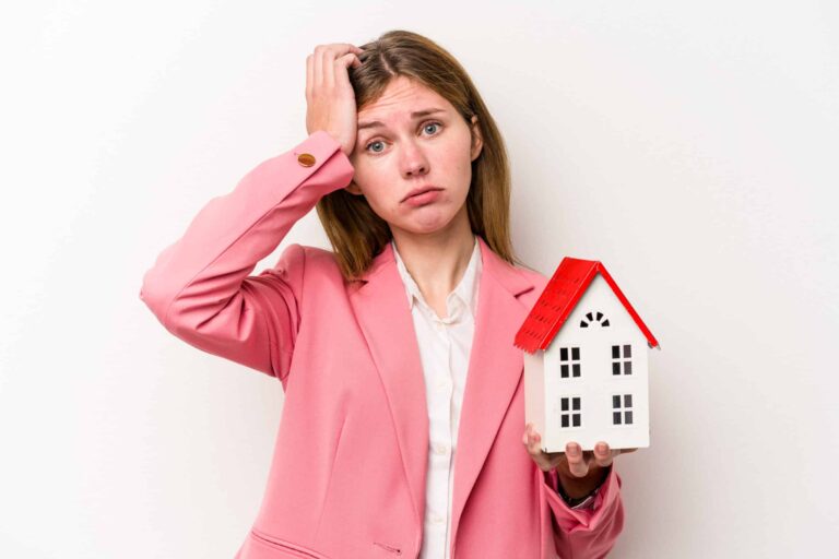 A woman in a pink blazer looks worried while holding a small model house with a red roof.