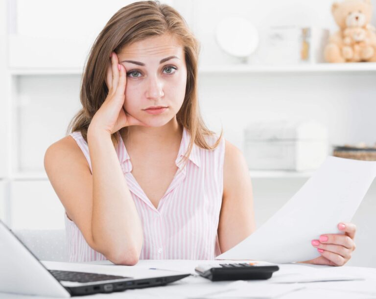 A woman looks tired while sitting at a desk with a laptop, a document in one hand, and a calculator nearby.
