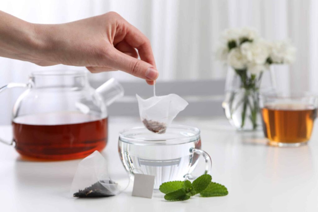 A hand places a tea bag into a clear glass cup filled with water. A teapot, a cup of tea, fresh mint, and white flowers are in the background.