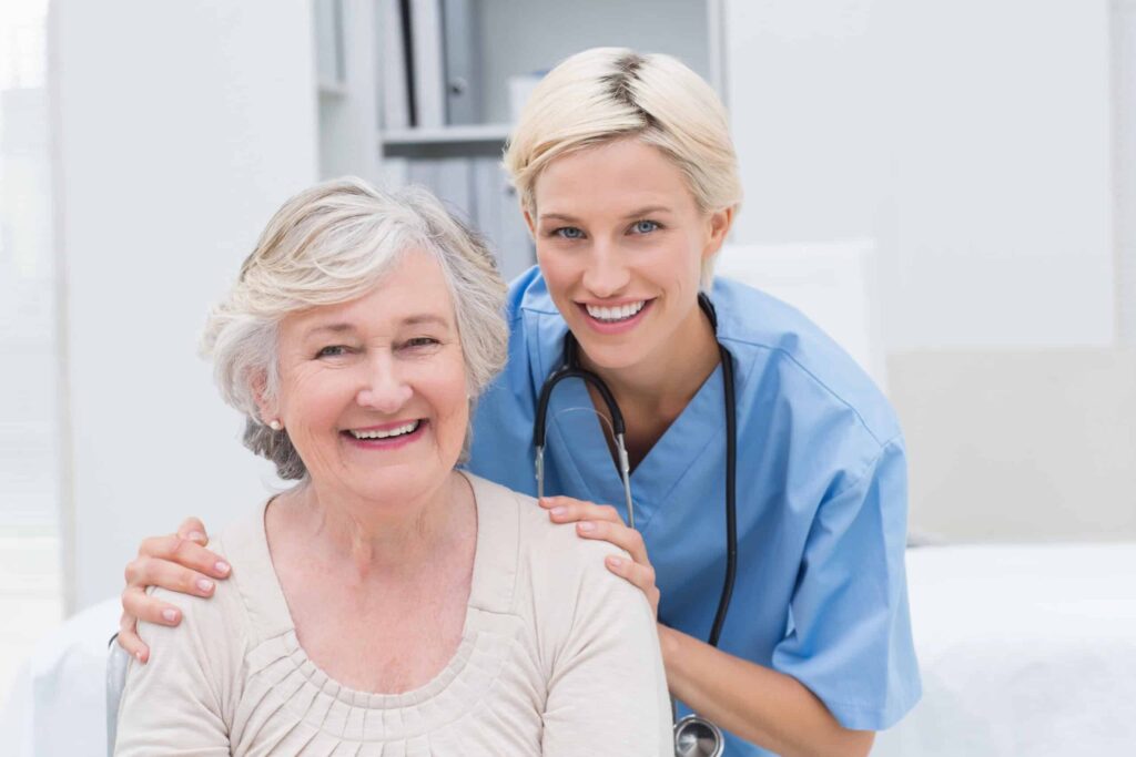 A smiling healthcare worker in blue scrubs with a stethoscope stands behind and gently touches an elderly woman seated and smiling in a room with medical equipment.