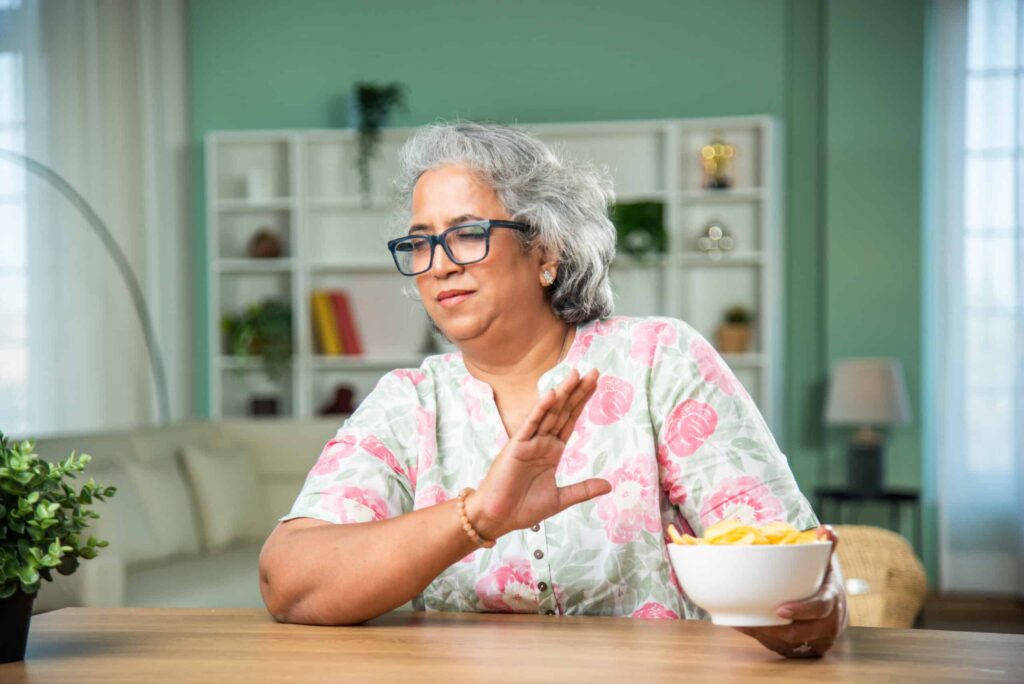 A woman in glasses and floral shirt holding a bowl of chips with one hand and gesturing to stop with the other in a living room.