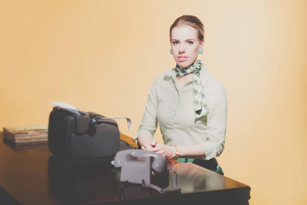 A woman with light hair sits at a desk with a vintage typewriter and telephone, wearing a light blouse, patterned scarf, and earrings.