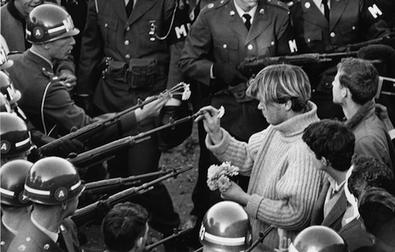 A man in a sweater places flowers into the rifle barrels held by several soldiers as others look on.