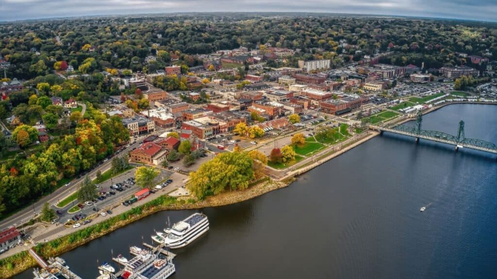 Aerial view of a riverside city showcasing a mix of tree-lined streets, buildings, and a marina with docked boats, set against a backdrop of a broad waterway and a bridge.