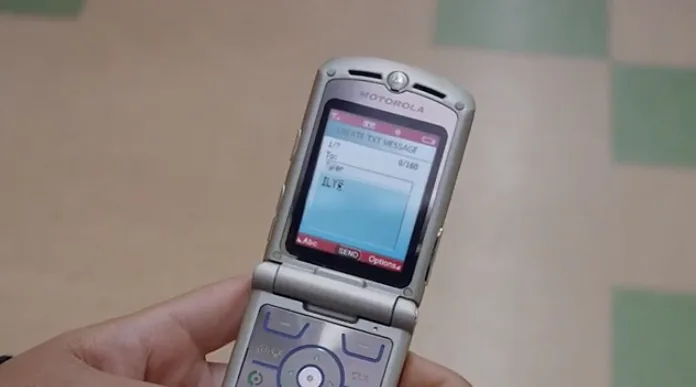 A person holding a Motorola flip phone displaying a text message draft on the screen. The phone has a silver exterior with a keypad visible. The background is slightly blurred.