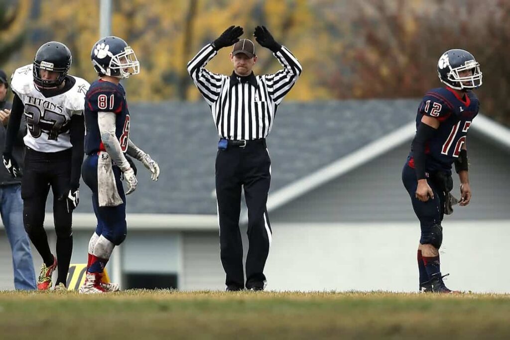 A football referee in a black and white striped uniform signals with raised arms, flanked by players in black and white, as well as blue and red uniforms, on a grassy field.