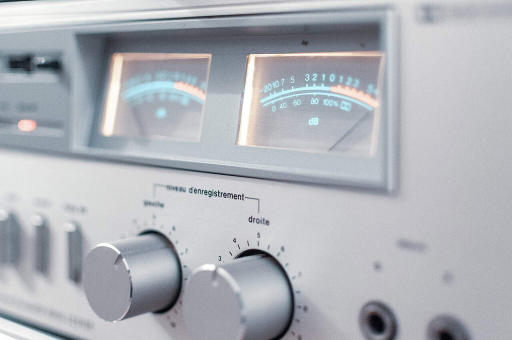 Close-up of a vintage audio receiver with dials and an analog level meter displaying signal levels. The receiver has a silver finish and labeled controls for recording.
