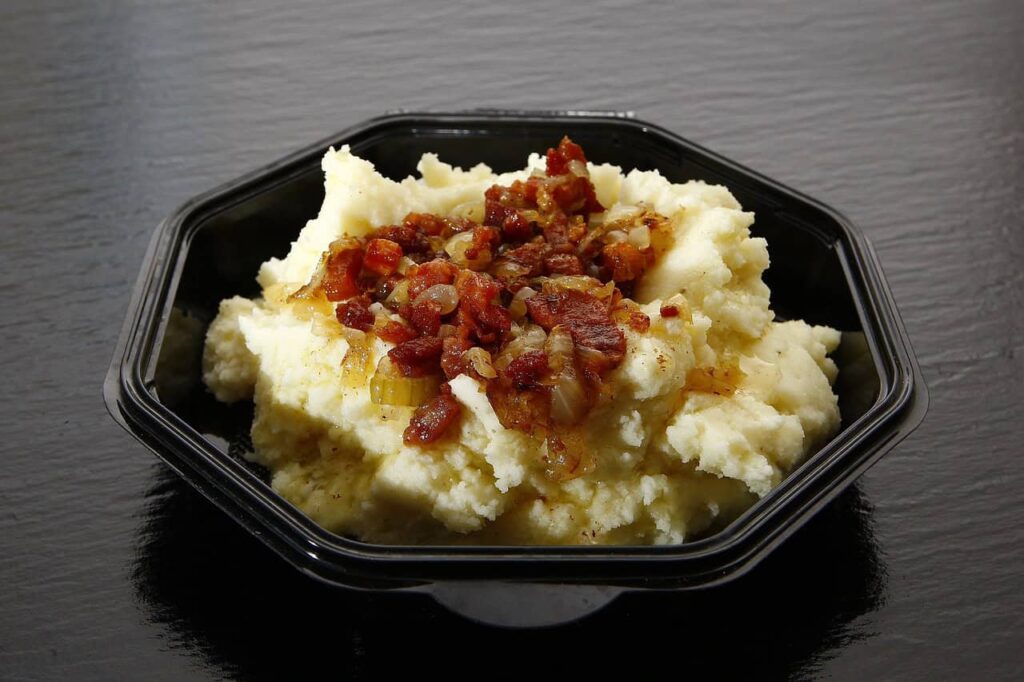 A black bowl containing mashed potatoes topped with chopped bacon and sautéed onions.