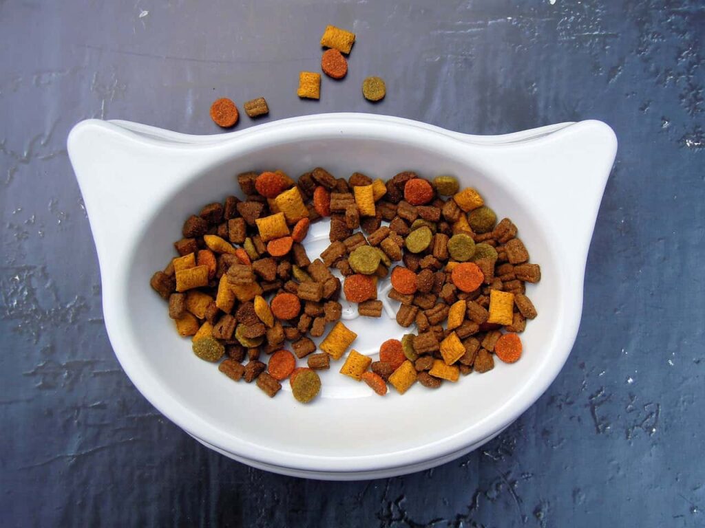 A white cat-shaped bowl filled with various colored dry cat food pellets on a gray textured surface.