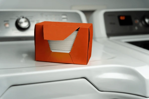 An orange box containing dryer sheets is placed on top of a white washing machine and dryer set.