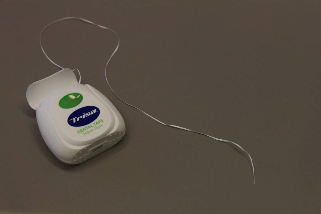 A white container of dental floss with an open lid and a piece of floss extending out. The container is labeled "Trisa Dental Tape.
