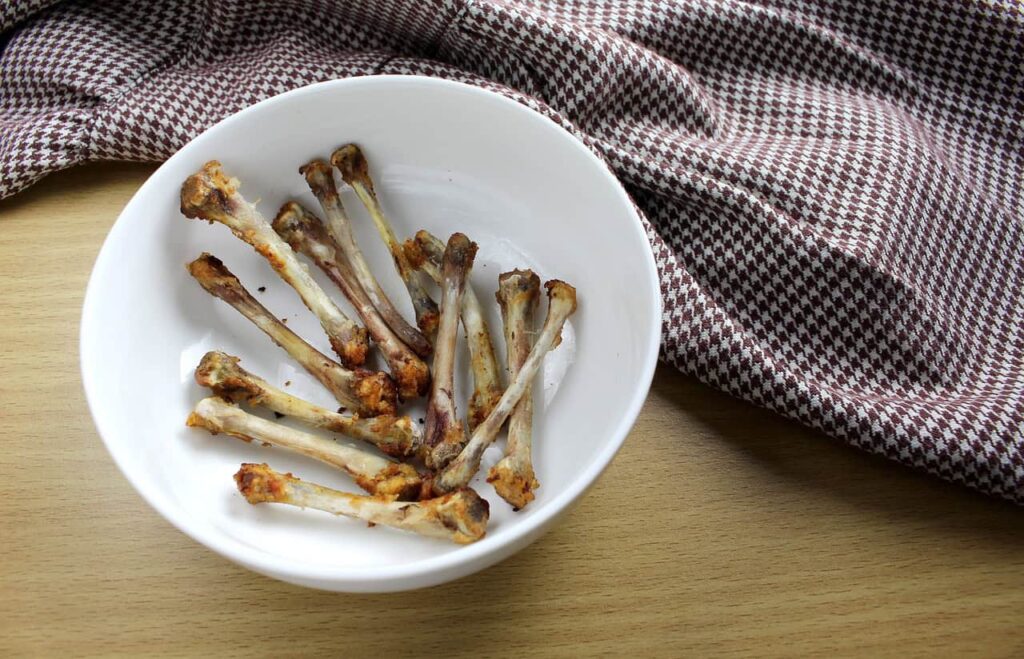 A white bowl containing multiple chicken bones sits on a wooden surface next to a brown and white checkered cloth.