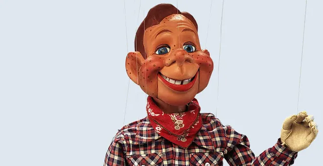 A puppet with a large smiling face, freckles, and blue eyes, wearing a red bandana and a plaid shirt, is shown against a plain background with strings attached to its head and hands.