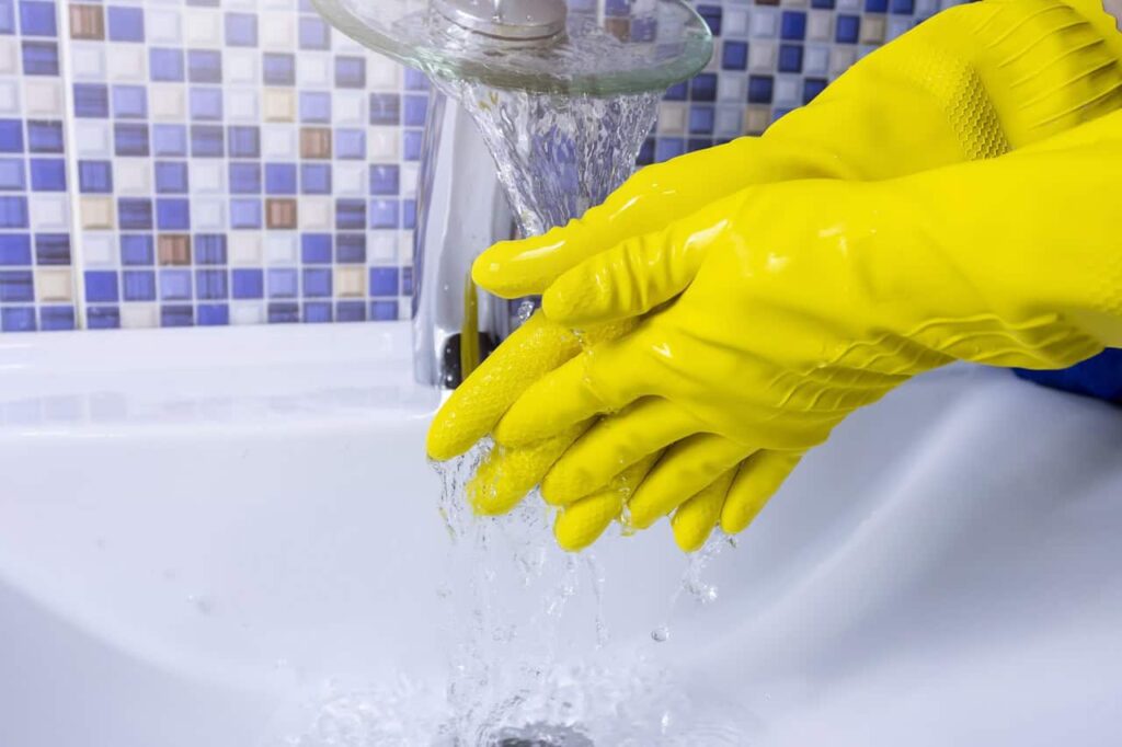 Yellow-gloved hands being washed under running water in a sink, with a blue and white tiled backsplash in the background.