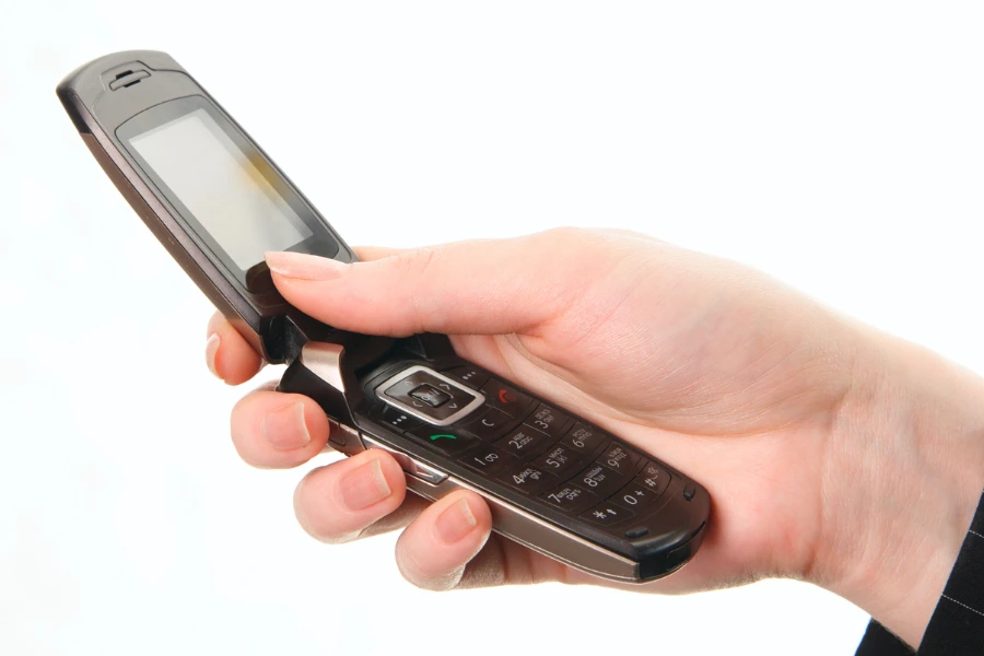 A person holds an open flip phone with an extended antenna in their right hand, displaying its keypad and small screen.