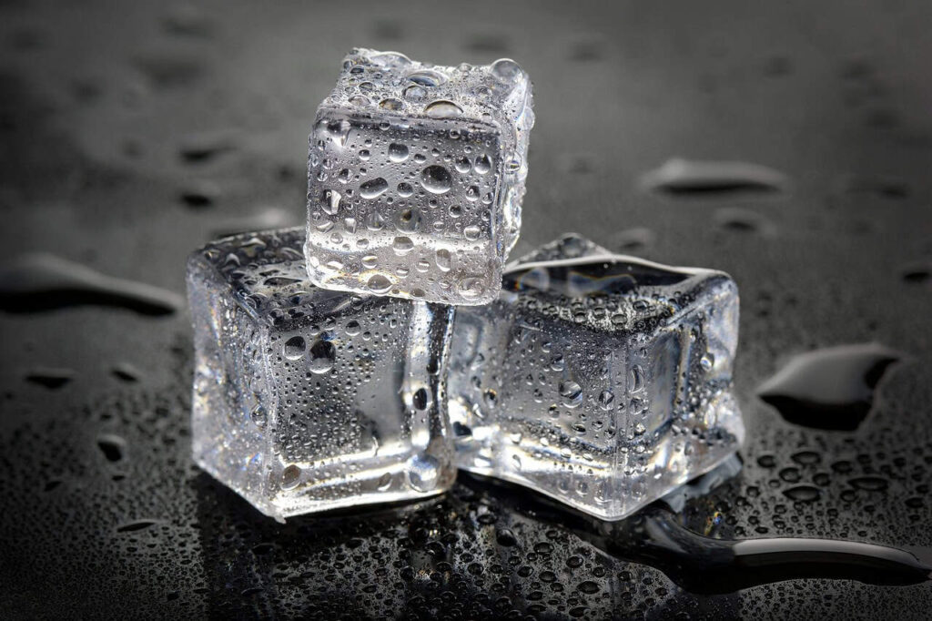 Three melting ice cubes with water droplets, stacked on a wet, reflective surface.