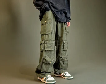 A person stands wearing an oversized black shirt, green cargo pants with multiple pockets, and green sneakers with white soles. The background is dark and plain.