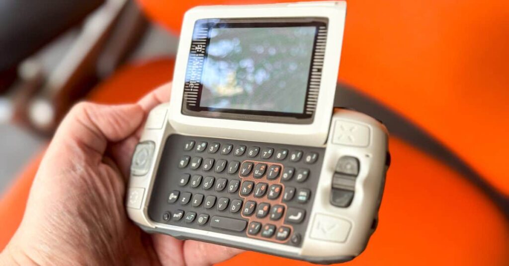 A person holds a vintage mobile handheld device with a flip-up screen and a full QWERTY keyboard against a blurred orange background.