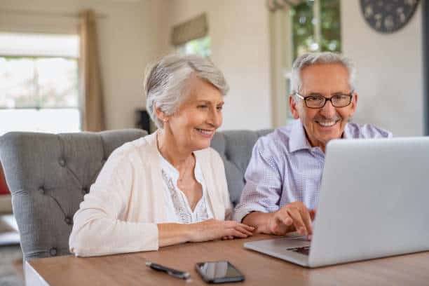 An elderly couple sits at a wooden table, smiling as they use a laptop. A smartphone and a pair of glasses lie on the table. The background features a window and a wall clock.