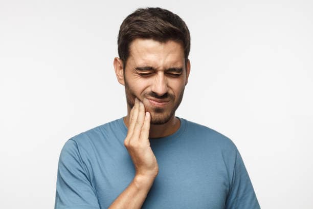 A man in a blue shirt holds his cheek with a pained expression, indicating dental discomfort or toothache.