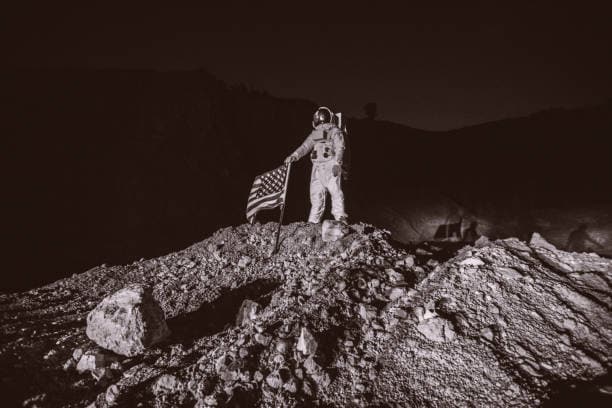 An astronaut in a space suit plants an American flag on a rocky, uneven surface in a barren landscape, under a dark sky.