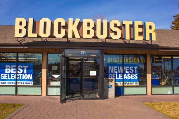 The storefront of a Blockbuster video rental store with signage advertising the best selection and newest releases. The entrance has double glass doors and the store's address is 211.