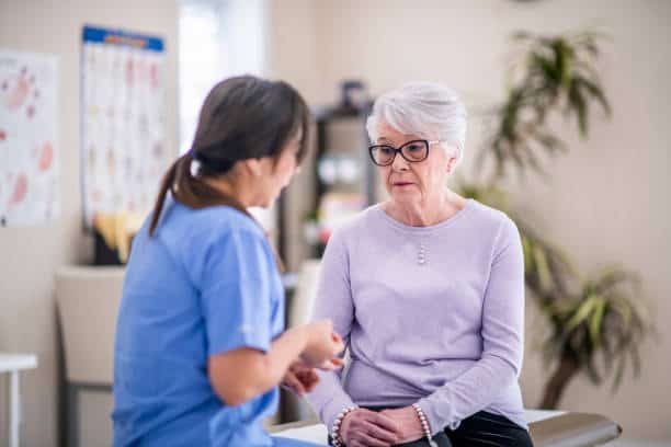 A nurse in blue scrubs talks to an elderly woman wearing glasses and a light purple sweater, who is seated on an examination table in a medical office.